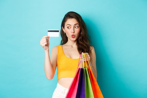 Imagem de mulher viciada em compras, mostrando seu cartão de crédito de plástico, segurando sacolas de compras, vestindo roupas de verão, em pé contra um fundo azul.