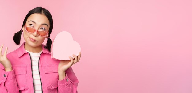 Imagem de namorada asiática estilosa em óculos de sol adivinhando o que está dentro da caixa de presente presente em forma de coração sobre fundo rosa