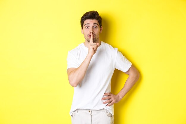 Imagem de um cara animado planejando uma surpresa, calando a câmera, pressionando o dedo contra os lábios em sinal de silêncio, em pé sobre um fundo amarelo