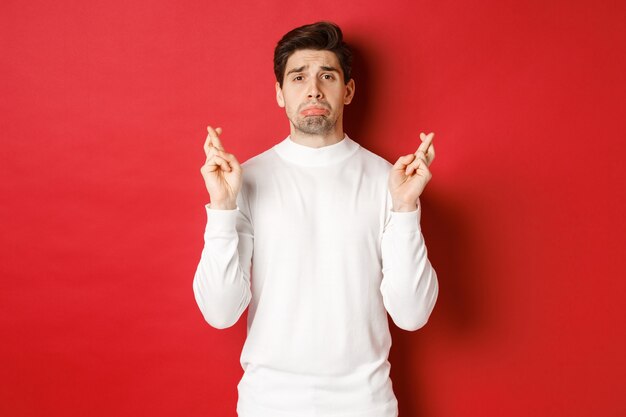 Imagem de um cara triste e sombrio em um suéter branco, chorando com os dedos cruzados, esperando por algo ou orando, em pé sobre um fundo vermelho