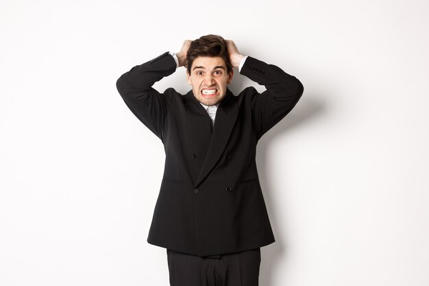 Imagem de um empresário frustrado e zangado em um terno preto, arrancando os cabelos da cabeça e fazendo uma careta de raiva, de pé tenso contra um fundo branco