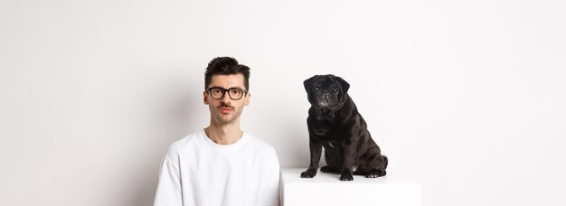 Foto grátis imagem de um hipster bonito em óculos sentado ao lado de um cachorro pug preto e fofo, ambos olhando para a câmera