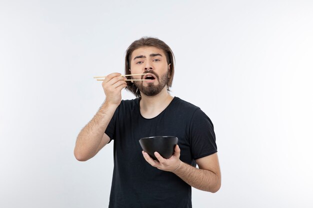 Imagem de um homem barbudo segurando uma tigela com pauzinhos sobre uma parede branca.