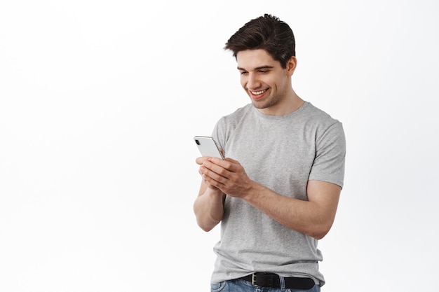 Imagem de um homem bonito escrevendo mensagem, conversando nas redes sociais e sorrindo, lendo a tela do celular, em pé sobre uma parede branca