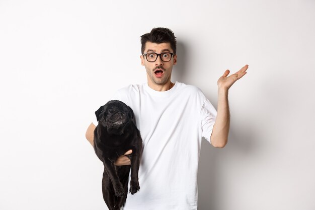 Imagem de um homem hippie confuso segurando cachorro e dando de ombros, não sei, levantando a mão intrigado, em pé com seu animal sobre um fundo branco