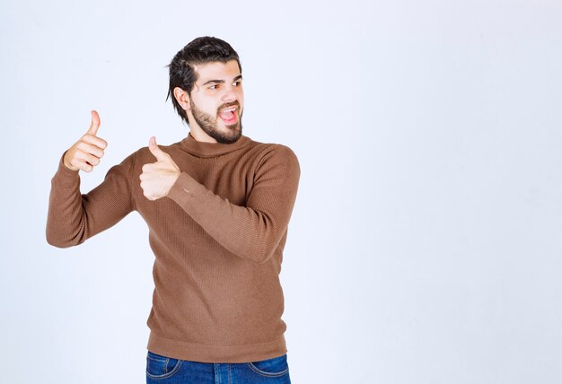 Imagem de um jovem atraente vestido com um suéter marrom em pé sobre um fundo branco mostrando um gesto de polegar para cima