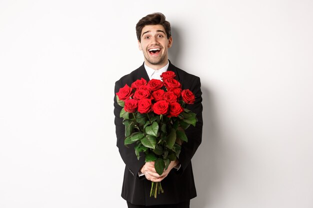Imagem de um lindo namorado em um terno preto, segurando um buquê de rosas vermelhas e sorrindo, estando em um encontro, em pé sobre um fundo branco