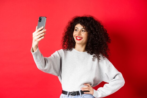 Imagem de uma blogueira elegante tomando selfie no smartphone, posando para uma foto no celular, em pé no fundo vermelho