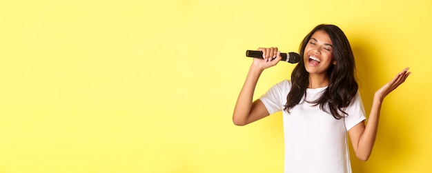 Foto grátis imagem de uma garota africano-americana despreocupada cantando no microfone sorrindo feliz se apresentando sobre ba amarelo