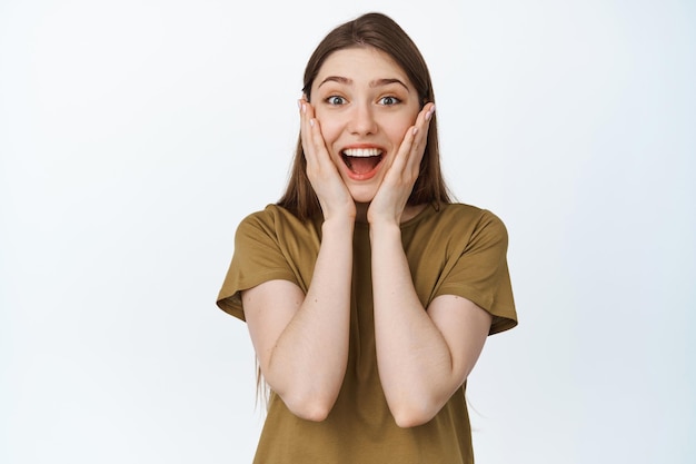 Imagem de uma garota animada e surpresa sorrindo, segurando as mãos no rosto emocionada, de pé na camiseta contra o fundo branco