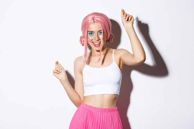 Imagem de uma garota atraente festeira com peruca rosa e maquiagem brilhante, se divertindo e comemorando o feriado, dançando feliz sobre fundo branco
