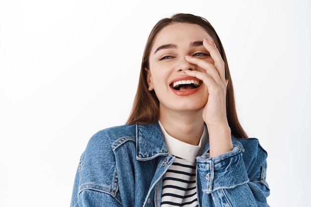 Foto grátis imagem de uma garota cândida rindo e parecendo feliz à parte no espaço da cópia do logotipo, tocando a pele natural sem maquiagem e sorrindo, em pé sobre um fundo branco