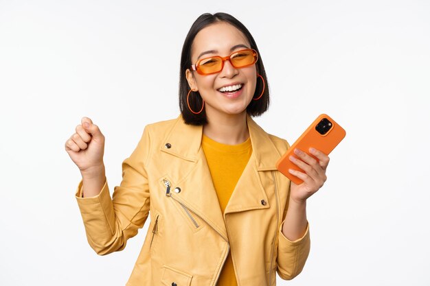 Imagem de uma garota coreana estilosa dançando com smartphone rindo feliz e sorrindo em pé sobre fundo branco