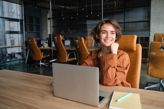 Foto grátis imagem de uma garota em seu local de trabalho, nômade digital, usando um laptop, sentada em um escritório, vestindo roupas casuais