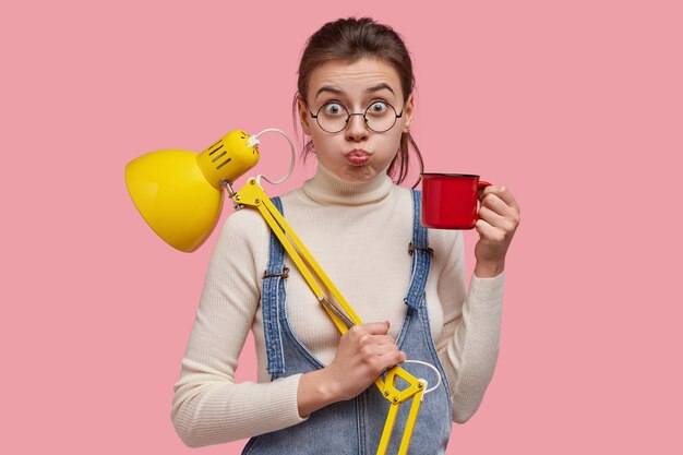 Imagem de uma jovem atraente assoando as bochechas, fazendo caretas, bebendo café ou chá, usa abajur para iluminar o ambiente, usa óculos redondos