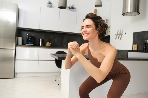 Imagem de uma jovem fazendo agachamentos em casa, exercitando-se em tapete de borracha em uma sala brilhante, vestindo activ