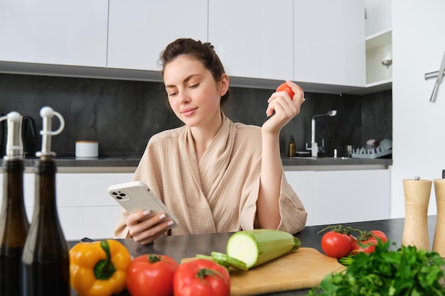 Imagem de uma jovem linda segurando tomate sentado na cozinha com uma tábua de cortar smartphone e