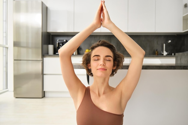 Imagem de uma jovem meditando levantando as mãos acima da cabeça praticando atenção plena em casa fazendo y