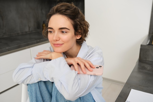 Imagem de uma jovem morena sentada sonhadora na cozinha sorrindo pensativa segurando o smartphone