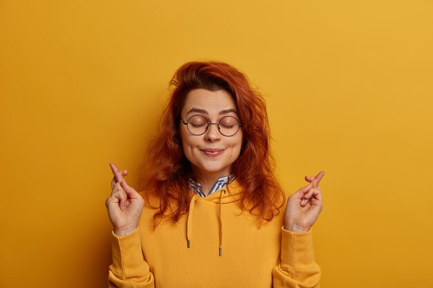 Imagem de uma jovem ruiva esperançosa de aparência agradável mantém os dedos cruzados, deseja boa sorte antes de passar no exame ou ir para uma entrevista de emprego, fecha os olhos