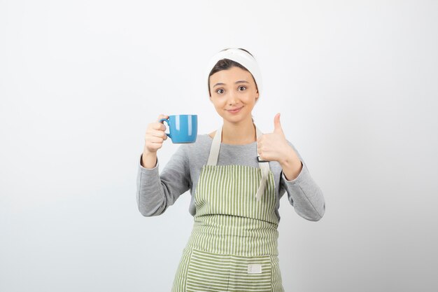 Imagem de uma jovem sorridente de avental com uma xícara azul aparecendo com o polegar para cima