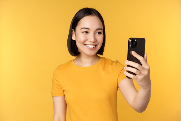 Imagem de uma linda garota asiática feliz conversando em vídeo falando no aplicativo de smartphone em pé contra o fundo amarelo