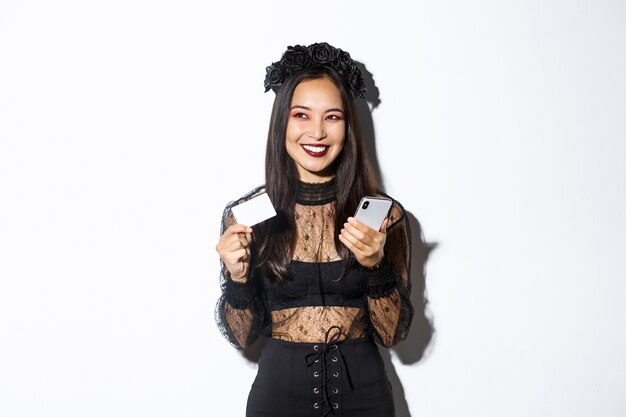 Imagem de uma linda mulher asiática em um vestido de renda gótica e grinalda preta, olhando de lado satisfeito e sorrindo, segurando o telefone celular com cartão de crédito.