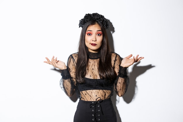 Imagem de uma menina asiática despreocupada e despreocupada, parecendo confusa, encolhendo os ombros em pé sobre um fundo branco com fantasia de halloween, vestida como uma bruxa má na festa.