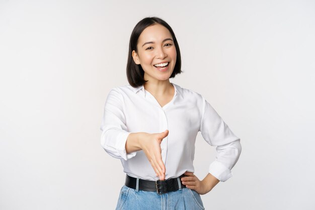 Imagem de uma mulher asiática confiante sorrindo estende a mão para o gesto de cumprimento de aperto de mão dizendo olá em pé sobre fundo branco