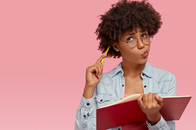 Imagem de uma mulher contemplativa de pele escura coçando a cabeça com um lápis, empurrando os lábios e olhando para cima com expressão pensativa e duvidosa, pensa no conteúdo criativo enquanto escreve a composição para a aula