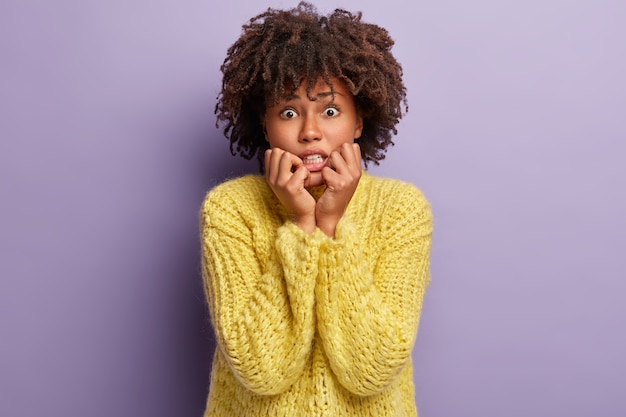 Imagem de uma mulher nervosa de pele escura que morde as unhas por causa da depressão, preocupada com a mágoa e a separação do namorado, tem corte de cabelo afro, usa um macacão amarelo, posa sozinha dentro de casa