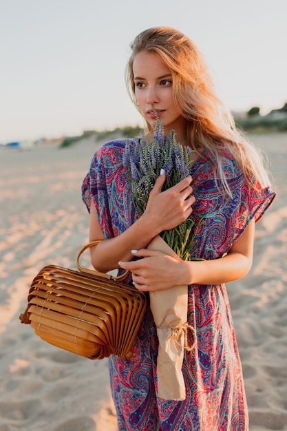 Imagem de verão ao ar livre da bela mulher loira romântica em vestido colorido, caminhando na praia com buquê de lavanda.