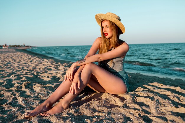 Imagem de verão ao ar livre de uma linda mulher loira com chapéu de palha, caminhando perto do mar.