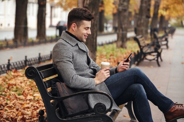 Imagem do homem morena sorridente no casaco e calça jeans, bebendo café para viagem e usando seu telefone celular, enquanto está sentado no banco do parque
