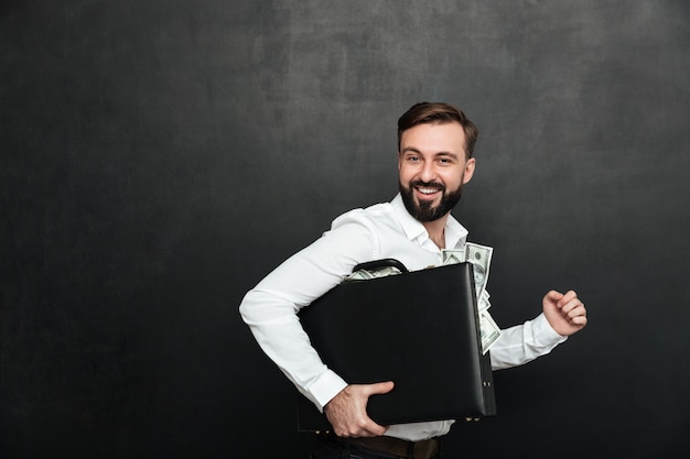 Imagem engraçada do homem de sorte, segurando a mala preta cheia de notas de dólar dentro, isolado sobre cinza escuro