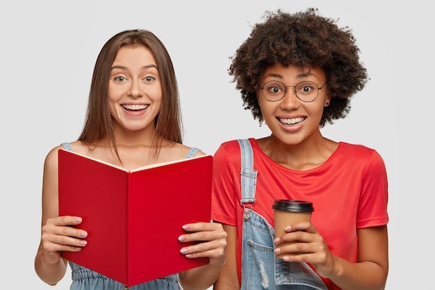 Foto grátis imagem horizontal de alegres dois alunos inter-raciais lendo livro, aprendendo material para o próximo exame, bebendo café quente para viagem