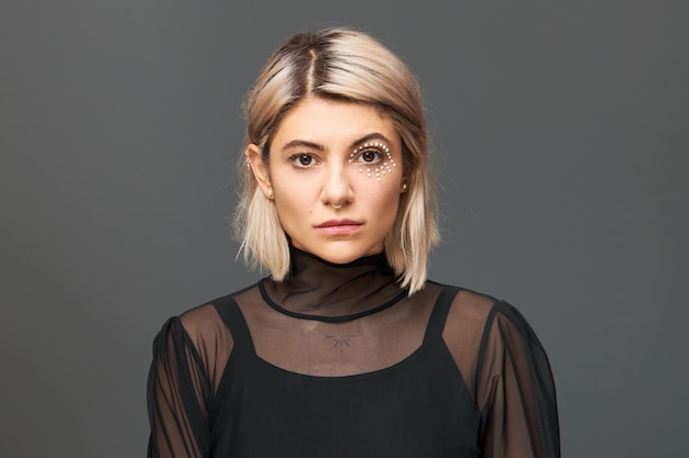 Imagem horizontal de incrível jovem mulher com penteado bob loiro e maquiagem artística com cristais brancos ao redor do olho posando isolado em uma blusa da moda, com olhar confiante