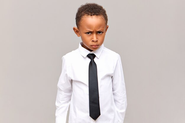 Imagem horizontal de um aluno de pele escura frustrado em uniforme escolar com uma expressão facial de raiva, carrancudo, zangado com um colega rude e valentão, pronto para se defender