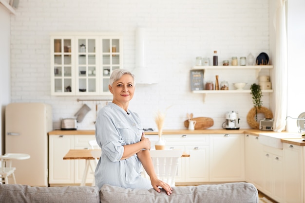 Imagem horizontal de uma linda dona de casa idosa de sessenta anos de idade descansando depois de limpar todos os quartos, com expressão facial alegre, em pé na sala de estar com cozinha