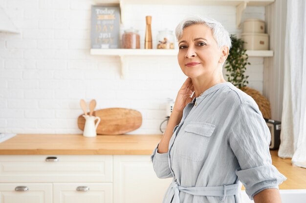 Imagem horizontal de uma linda mulher idosa madura usando um elegante vestido azul posando em um ambiente fechado, com um balcão de cozinha e utensílios limpos e aconchegantes