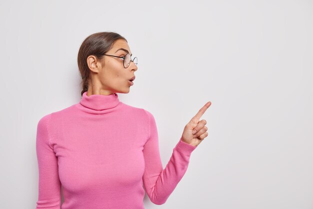 Imagem horizontal de uma mulher chocada e surpresa que reage a notícias incríveis usando vidros transparentes e pontas de gola rolê rosa casuais em um espaço de cópia em branco anuncia algo isolado sobre uma parede branca