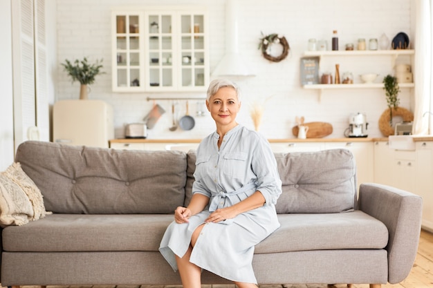 Imagem interna de atraente e elegante mulher aposentada caucasiana com cabelo curto cinza usando um elegante vestido azul, sentado no sofá em pose relaxada, olhando com um sorriso calmo e alegre. Pessoas e idade