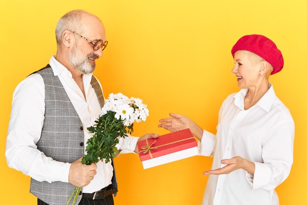 Imagem isolada da bela senhora madura europeia recebendo uma caixa de doces e flores do campo de seu namorado idoso em roupas elegantes e óculos. Homem sênior tímido fazendo um presente de aniversário para a esposa