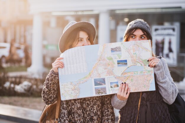 Imagem positiva engraçada de mulheres elegantes na rua ensolarada se divertindo na cidade, se escondendo atrás do mapa da cidade. Viajar juntos, melhores amigos, se perder na cidade grande, verdadeiras emoções.