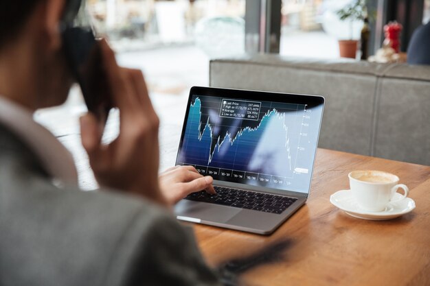 Imagem recortada de empresário sentado à mesa no café e analisando indicadores no computador portátil enquanto fala pelo smartphone