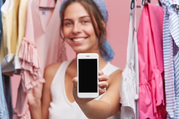 Imagem recortada de fêmea jovem tendo sorriso agradável em pé perto de prateleira com roupas, segurando o celular moderno na mão com tela em branco. Pessoas, tecnologias modernas e conceito de moda