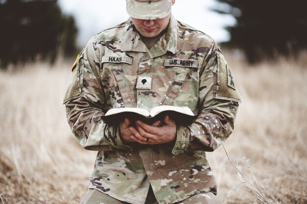 Imagem superficial de um jovem soldado ajoelhado em uma grama seca enquanto lia a Bíblia