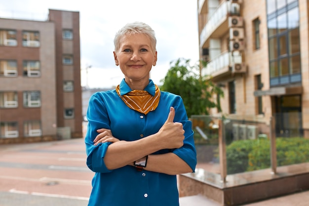 Imagem urbana ao ar livre de mulher madura confiante e bem-sucedida em roupas elegantes, posando em uma rua vazia com um edifício moderno de vários andares no fundo, com uma aparência positiva, fazendo um gesto de polegar para cima