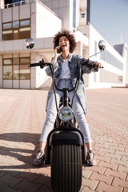 Imagem vertical da mulher encaracolada feliz sentado na moto moderna