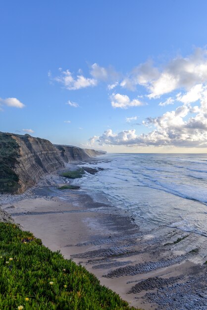 Imagem vertical da praia rodeada pelo mar e falésias ao sol e céu nublado